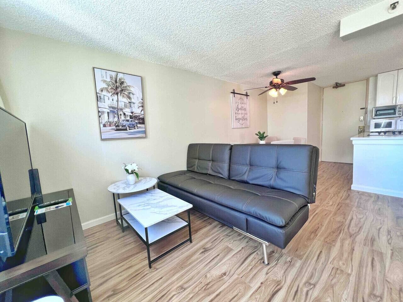 A living room with a dark leather sofa, a small round table, a flat-screen TV, and a ceiling fan.