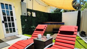 A sunlit outdoor seating area with red lounge chairs and a small table, surrounded by a neatly trimmed hedge and a wooden deck.