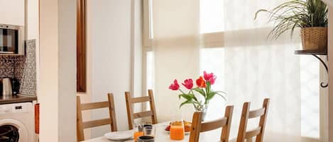 A dining area with a table set for two, chairs, a vase with flowers, and a window with sheer curtains.