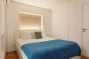 A bedroom with a bed, a bedside table, a window with blinds, and a door. - Alfama River Light | Sunlit Views & AC (Lisbon)