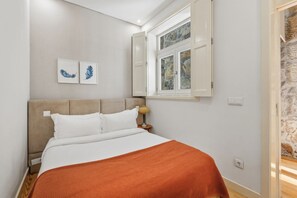 A bedroom with a bed, a nightstand, a lamp, a window with open shutters, and a wall-mounted light switch. - Recanto do Porto with Private Patio and AC (Porto)