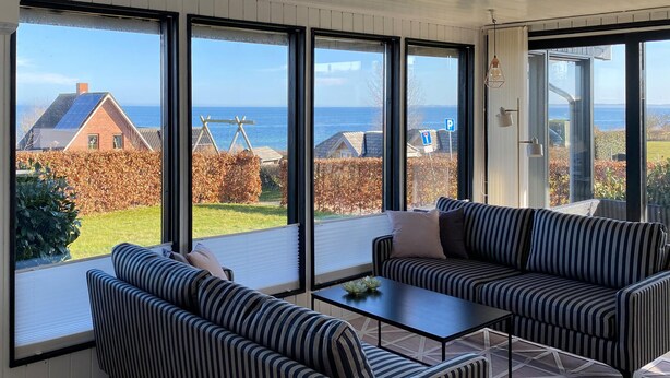 Quarto iluminado pelo sol, com grandes janelas, um sofá listrado, uma mesa de centro e vista para o mar e casas vizinhas.