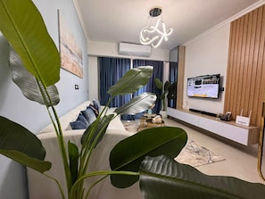 Ein modernes Wohnzimmer mit einer Couch, großen Grünpflanzen, einem Fernseher und einer Klimaanlage.