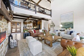 A spacious living area with a fireplace, a high ceiling with exposed beams, a balcony, and a large painting on the wall. - Views, Pickleball, Hottub, Gameroom, EV, Multi-Fam (Broken Bow)