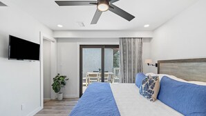 Une chambre à coucher avec un lit, un ventilateur de plafond, une télévision, une plante en pot et une vue sur l’extérieur.