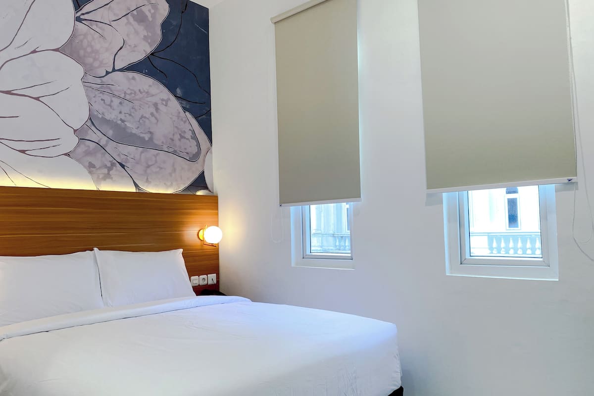 A hotel room with a bed, a large floral artwork on the wall, two windows with blinds, and a nightstand with a lamp.