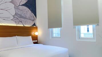 Habitación de hotel con cama, un cuadro grande con motivos florales en la pared, dos ventanas con persianas y una mesita de noche con lámpara.