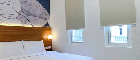 A hotel room with a bed, a large floral artwork on the wall, two windows with blinds, and a nightstand with a lamp.