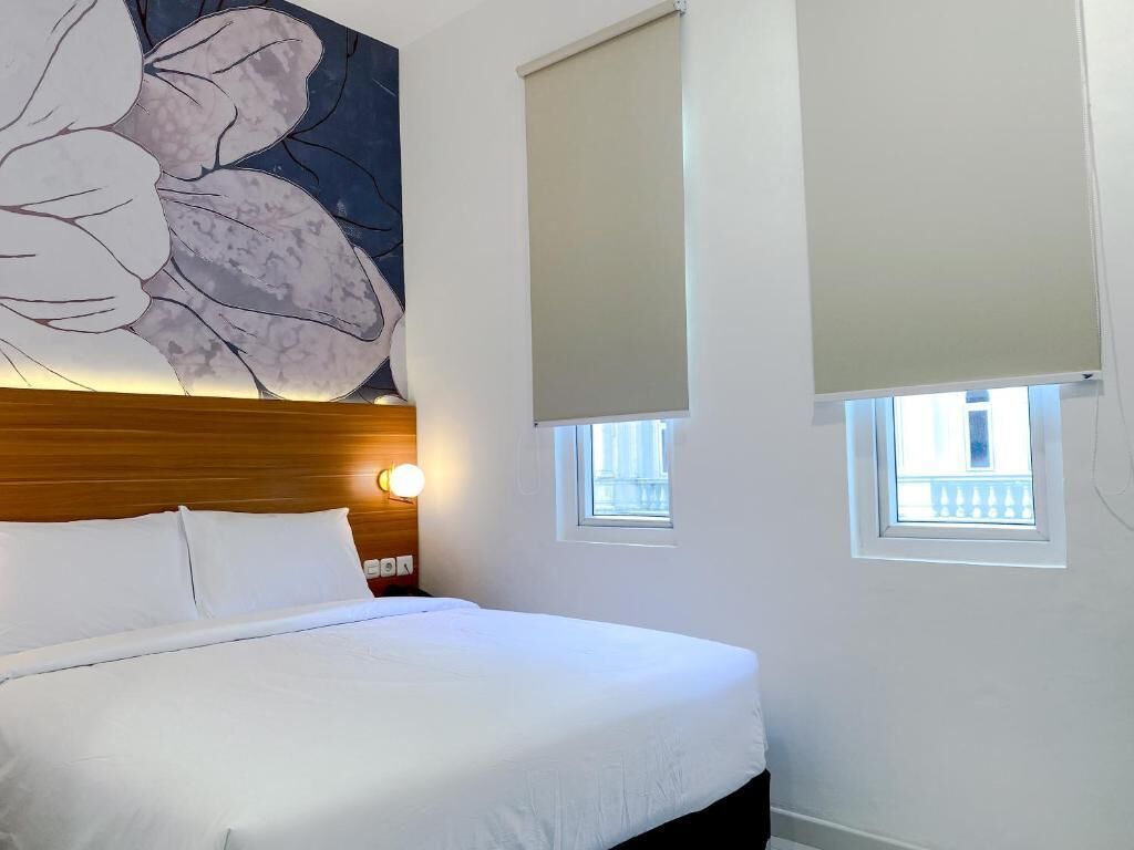 A hotel room with a large bed, a headboard with a light fixture, two windows with blinds, and a wall-mounted artwork.