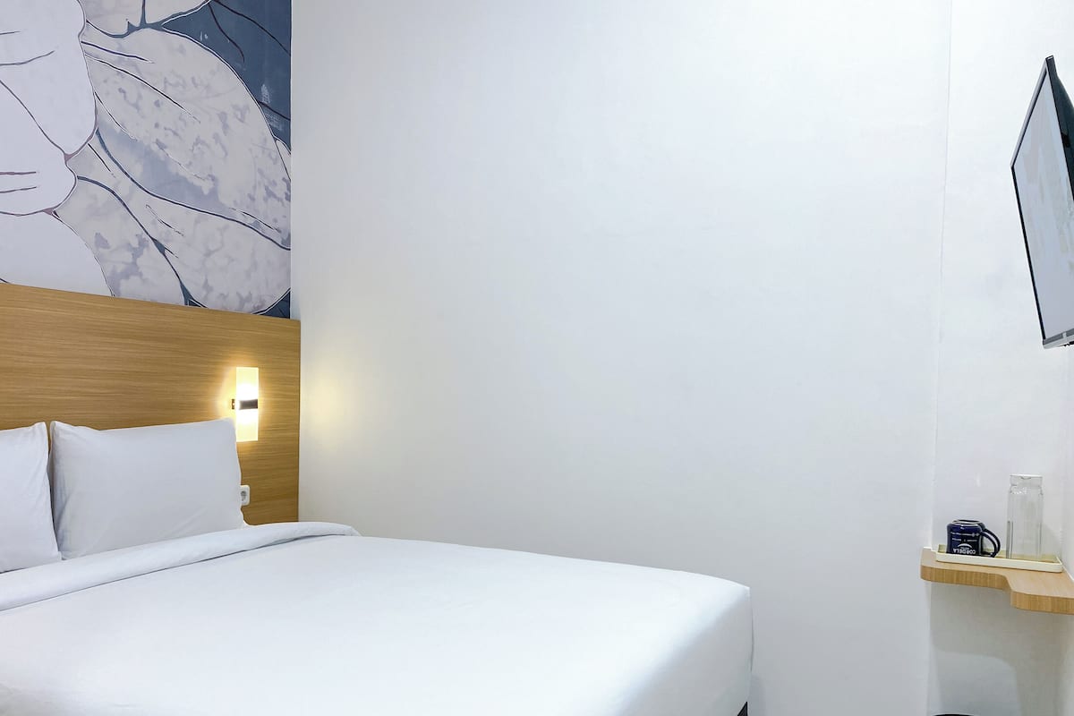 A hotel room with a bed, a headboard, a wall-mounted light, a shelf with a mug, and a trash bin.