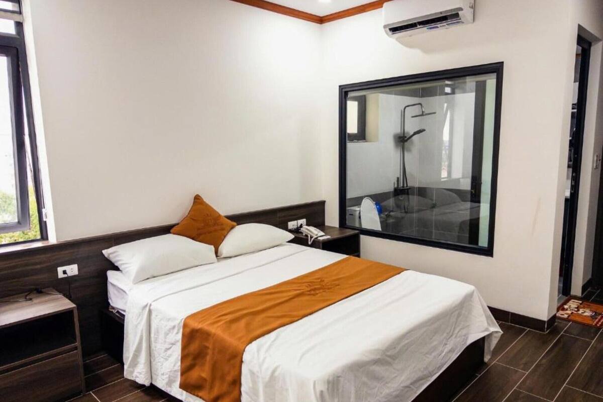 A hotel room with a bed, a nightstand, a bedside table, a window, and a bathroom visible through a glass wall.