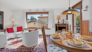 Ein Zimmer mit Esstisch, gedecktem Tisch, einer Couch mit roten Kissen, einem gemusterten Teppich und Blick auf ein Gebäude.