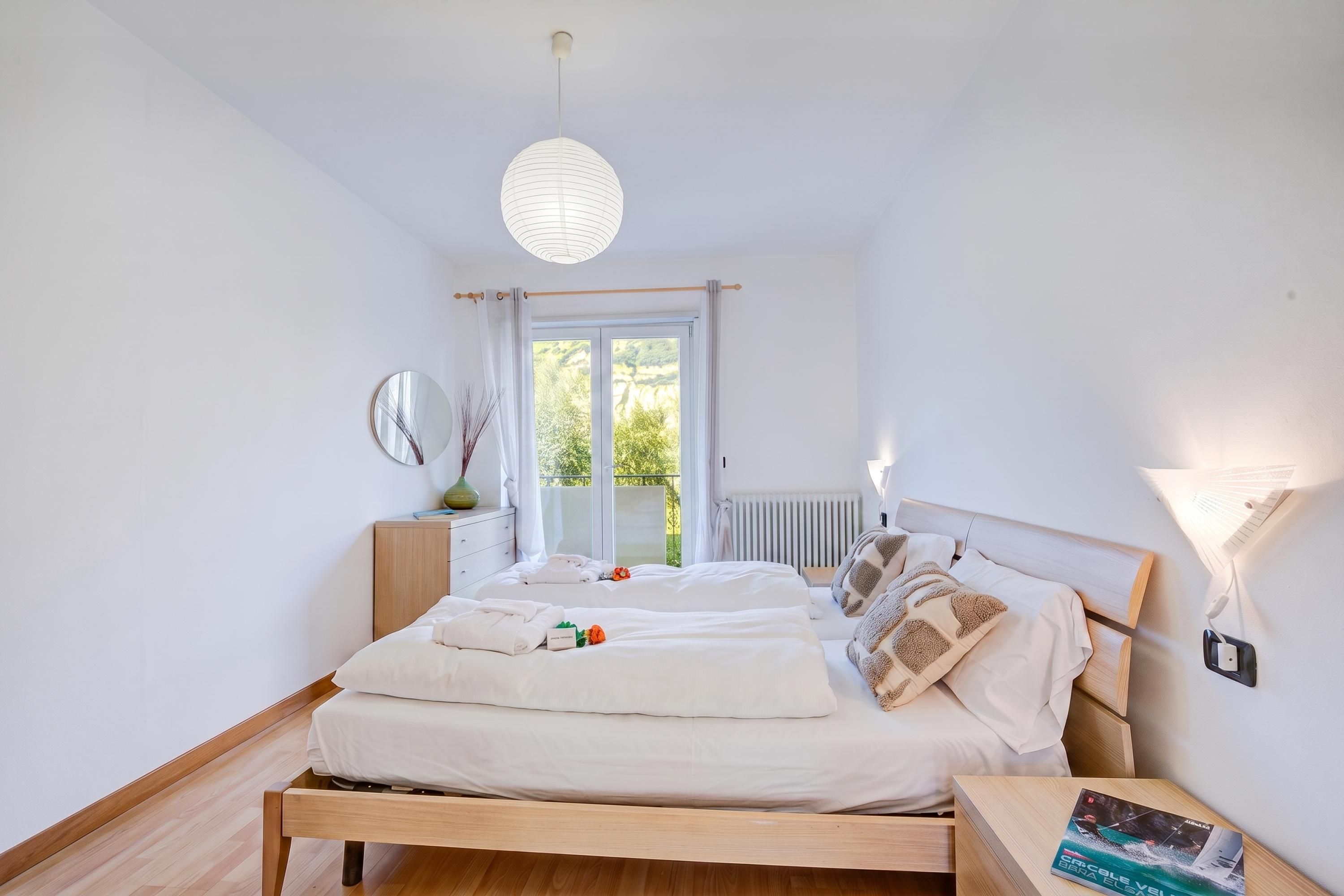 Quarto com cama de madeira, mesa de cabeceira, espelho e vista para o verde lá fora.