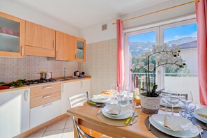 A modern kitchen with wooden cabinets, a dining table set for a meal, and a view of the outdoors through a window. - Agritur Laura- Essenza di rosa 2 with Shared Garden, Torbole sul Garda, Italy (Torbole sul Garda)