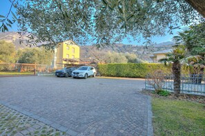 Ein gepflasterter Weg führt zu einem Gebäude, vor dem zwei Autos geparkt sind, umgeben von Grünflächen mit einer Bergkulisse im Hintergrund.