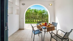 Uma área de refeições com mesa e cadeiras, vista para o verde através de um arco, e chão de azulejos.