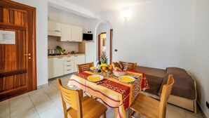 Uma área de refeições com uma mesa posta para dois, um sofá e uma cozinha ao fundo.