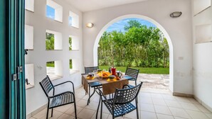 Uma área de refeições com uma mesa posta para quatro pessoas, rodeada por cadeiras, com vista para um jardim através de um arco.
