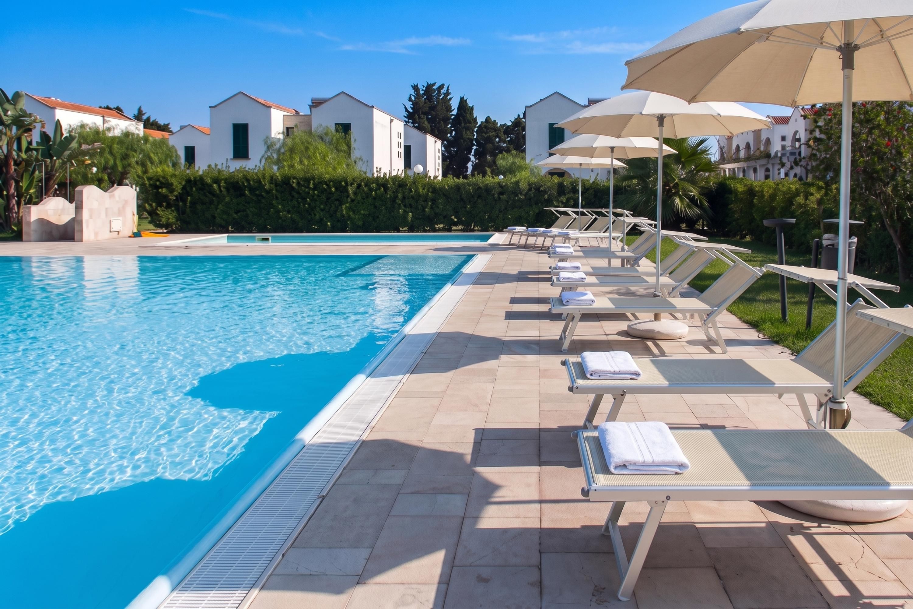 A poolside area with lounge chairs and umbrellas, a clear blue pool, and modern buildings in the background.