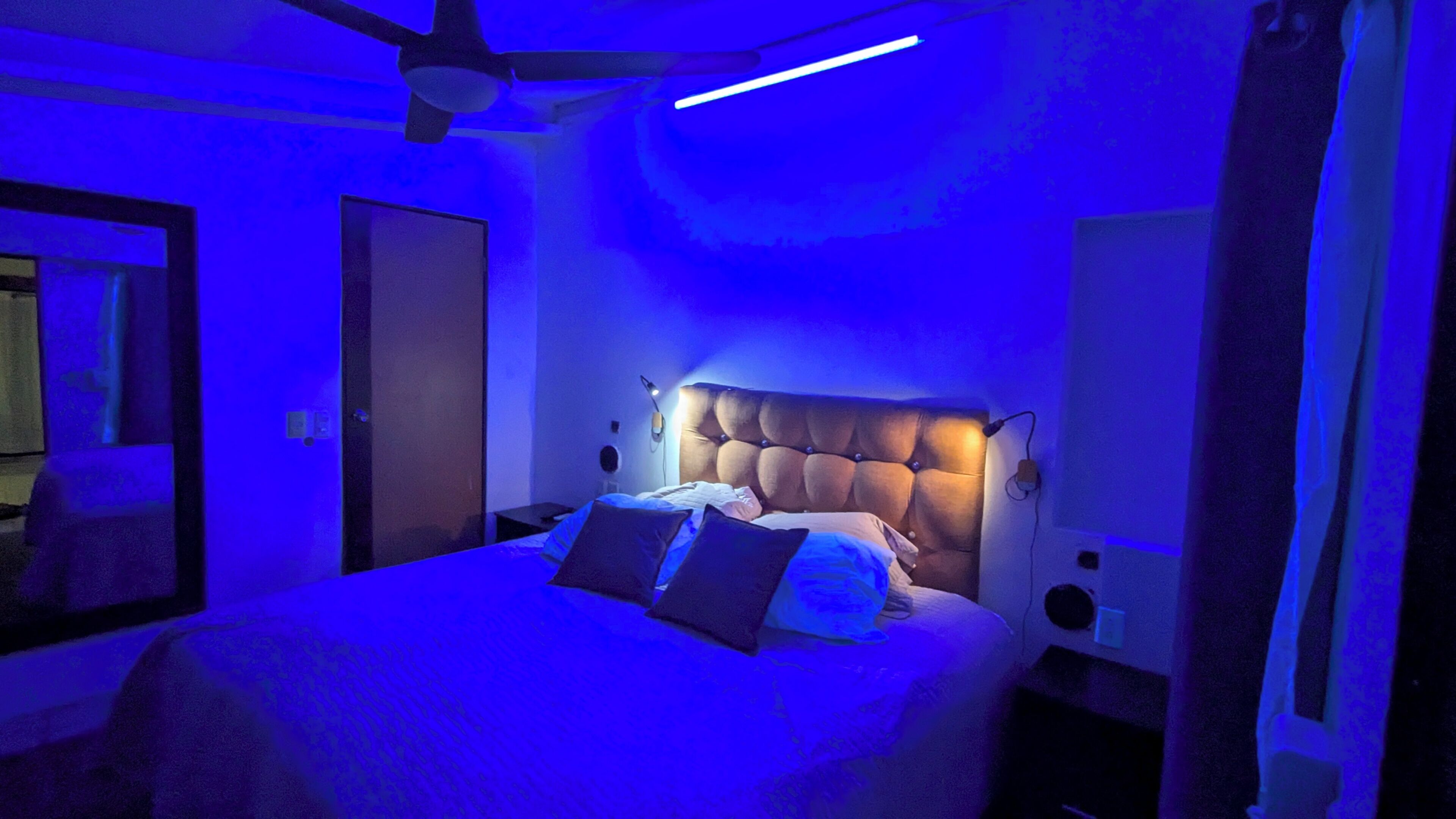 Une chambre à coucher avec une tête de lit capitonnée, un lit avec des oreillers, une table de chevet, un ventilateur de plafond et une lampe fixée au mur.