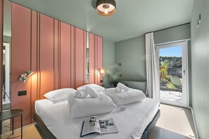 Ein Hotelzimmer mit Bett, Nachttisch, einer Zeitschrift, einer Wandleuchte und Blick ins Freie.