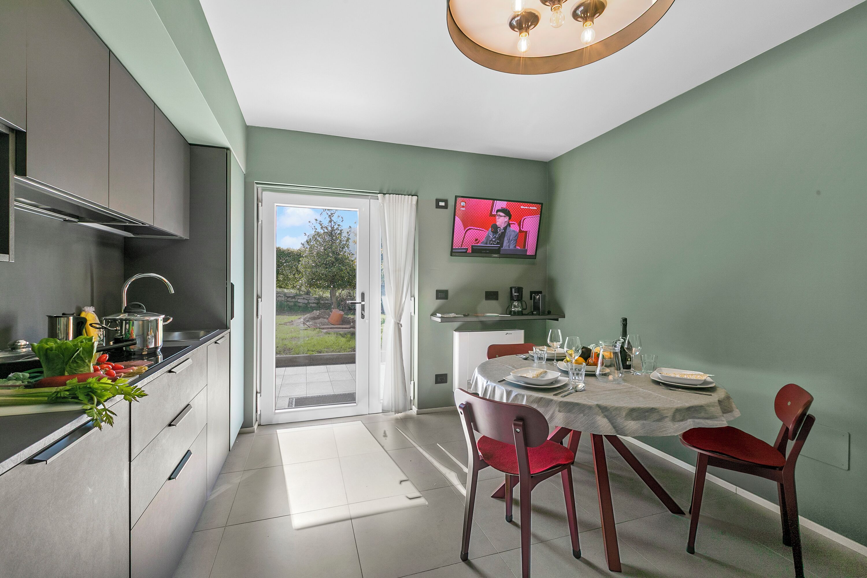 A modern kitchen with a dining area, a TV mounted on the wall, and a sliding glass door leading to an outdoor area.