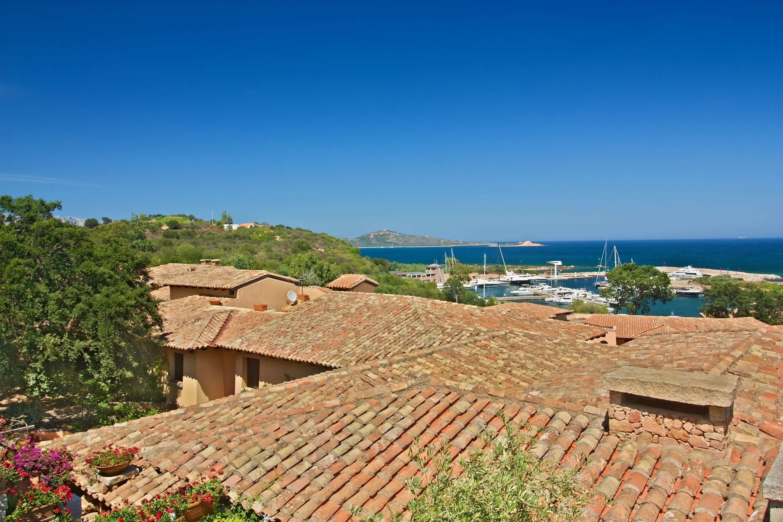 A coastal village with terracotta-roofed buildings, a marina with boats, and a clear blue sky.