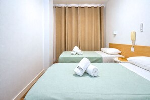 Una habitación de hotel con dos camas, cortinas color beige, una mesita de noche con una lámpara y un teléfono en la pared.