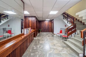 Zona de recepción con mostrador de madera, sillas rojas, suelo de mármol y una escalera con barandillas de madera.