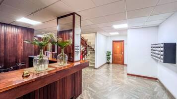 Zona de recepción con mostrador de madera, un espejo grande y dos jarrones con flores. Se ve una escalera al fondo que conduce a otro nivel.