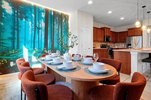 Zona pranzo con tavolo rotondo apparecchiato per sei persone, circondato da sedie arancioni, un murale a tema bosco e una cucina sullo sfondo.