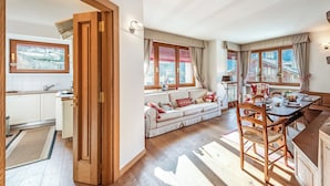 Eine moderne Küche mit Holzschränken, ein Essbereich mit Holztisch und -stühlen sowie eine gemütliche Sitzecke mit Sofa und Kissen.