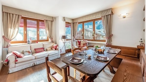 Ein gemütlicher Essbereich mit einem Holztisch, gedeckt für vier Personen, einer Couch mit roten Kissen und großen Fenstern mit Vorhängen.