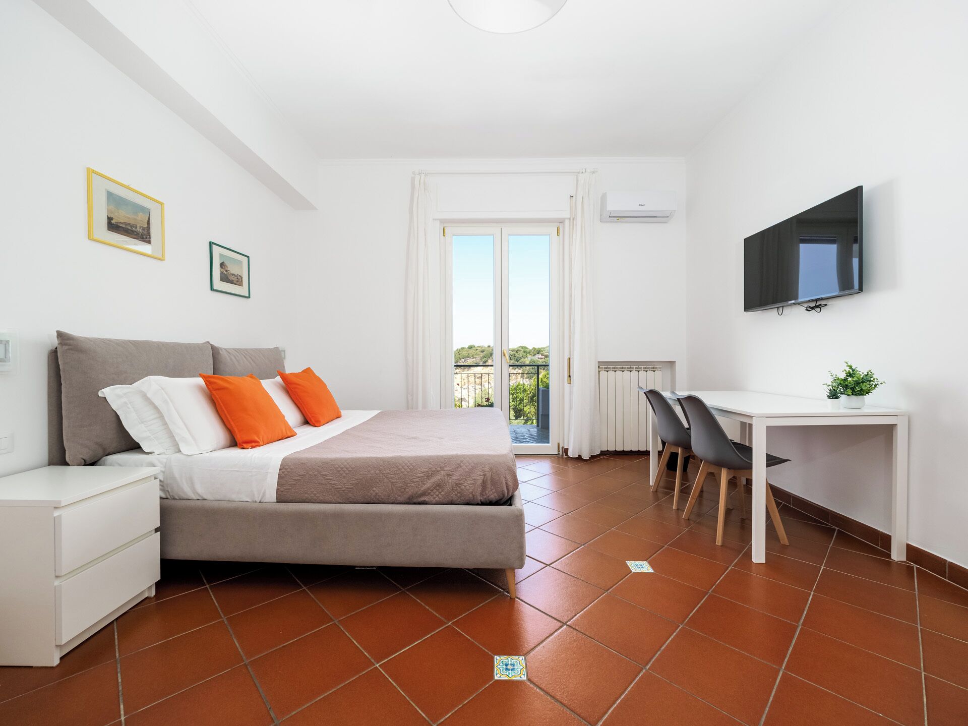 A bedroom with a bed, a desk, a chair, a TV, and a balcony with a view.