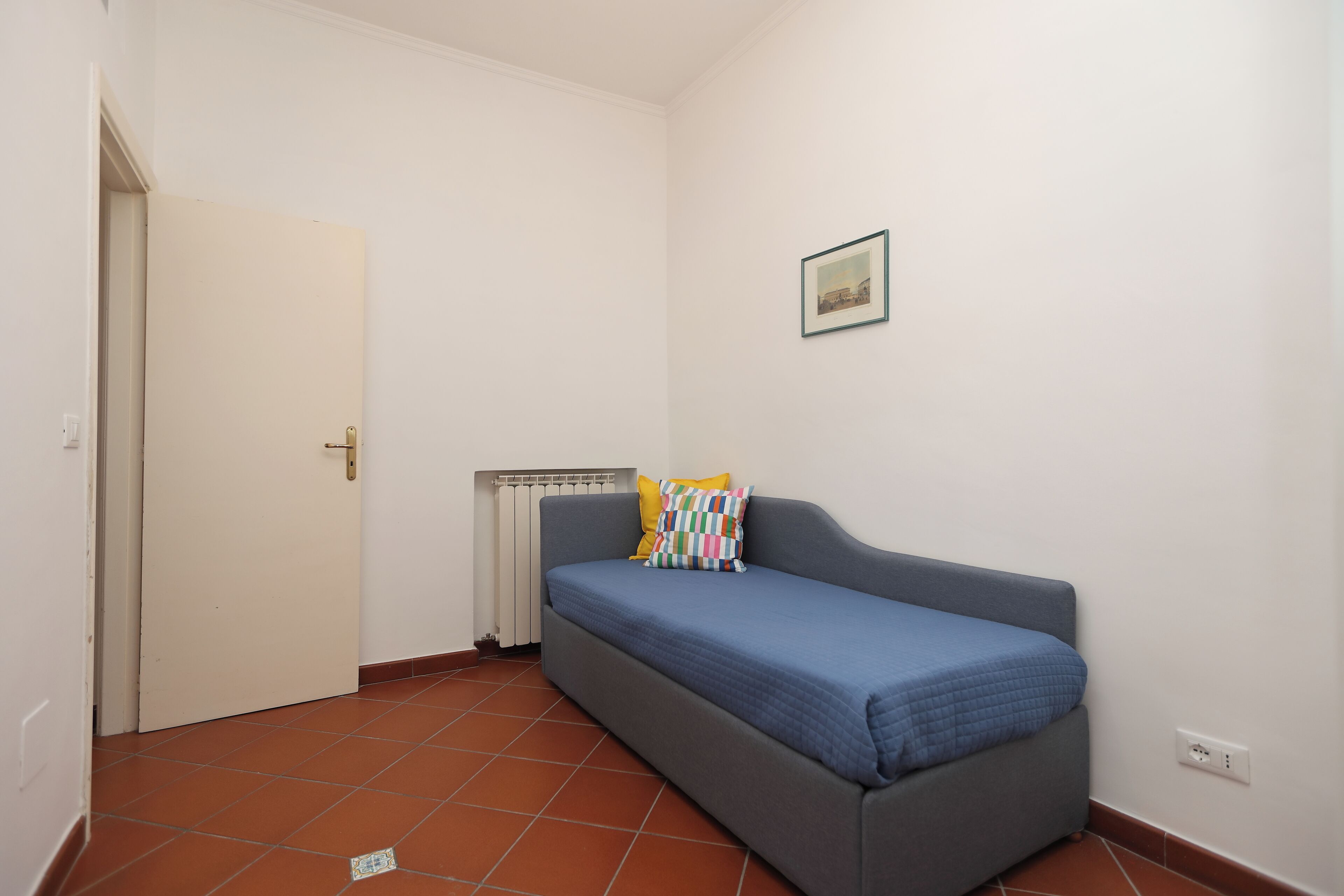 Una habitación pequeña con una cama individual, un radiador, una puerta blanca y un cuadro colgado en la pared.