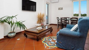 A living room with a TV, a dining table, chairs, a blue armchair, a coffee table, a potted plant, and a patterned rug. - Vervece House (Massa Lubrense)