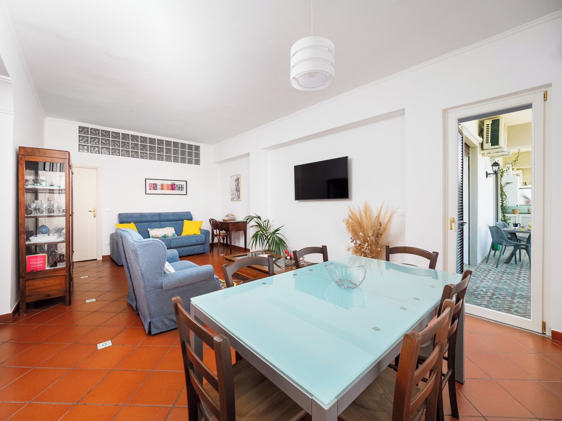 Un salon avec un canapé bleu, une table à manger avec des chaises, un téléviseur fixé au mur et une porte donnant sur un balcon.