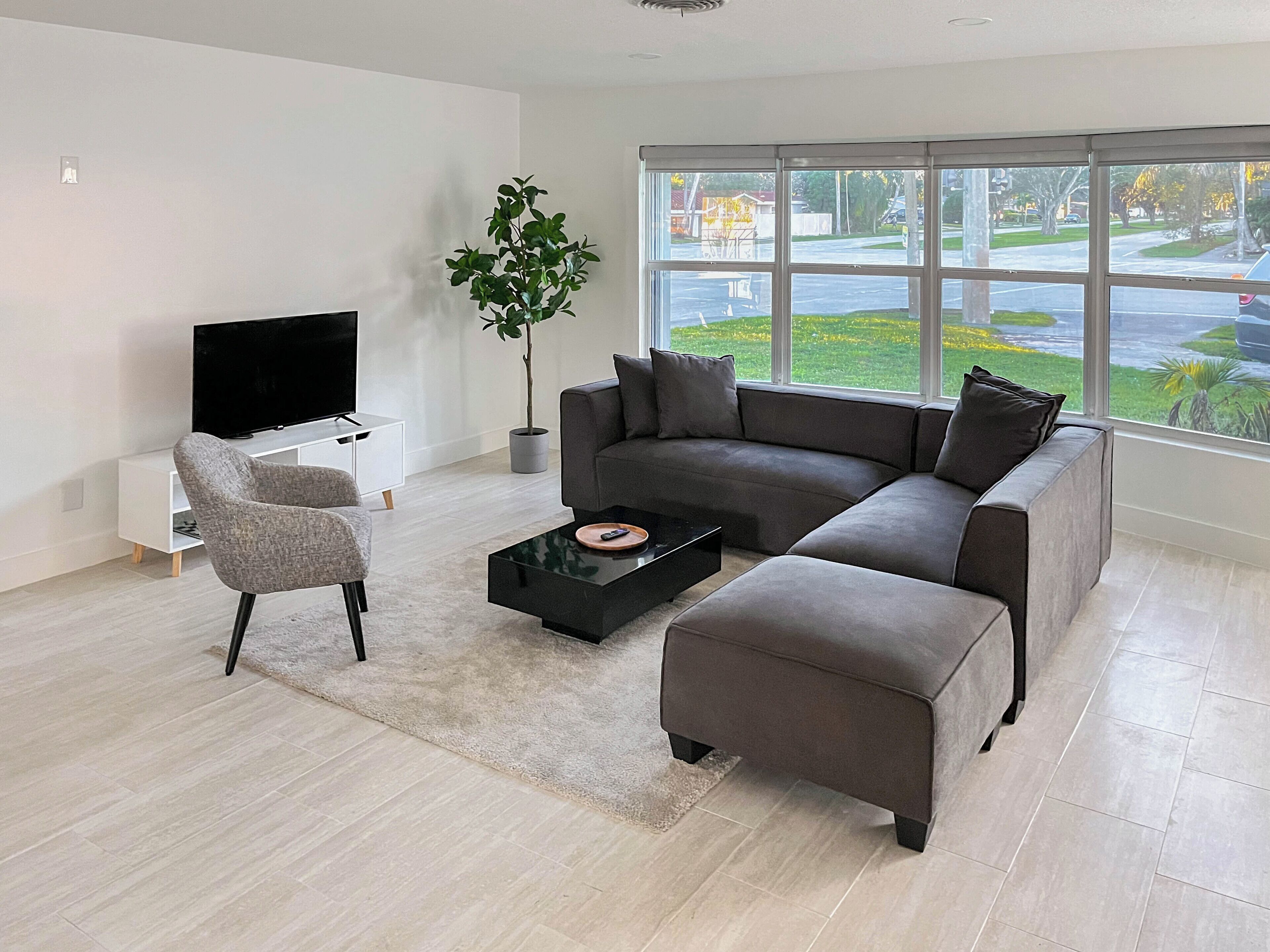 A modern living room with a sectional sofa, a TV, a coffee table, and a large window with a view of a street.