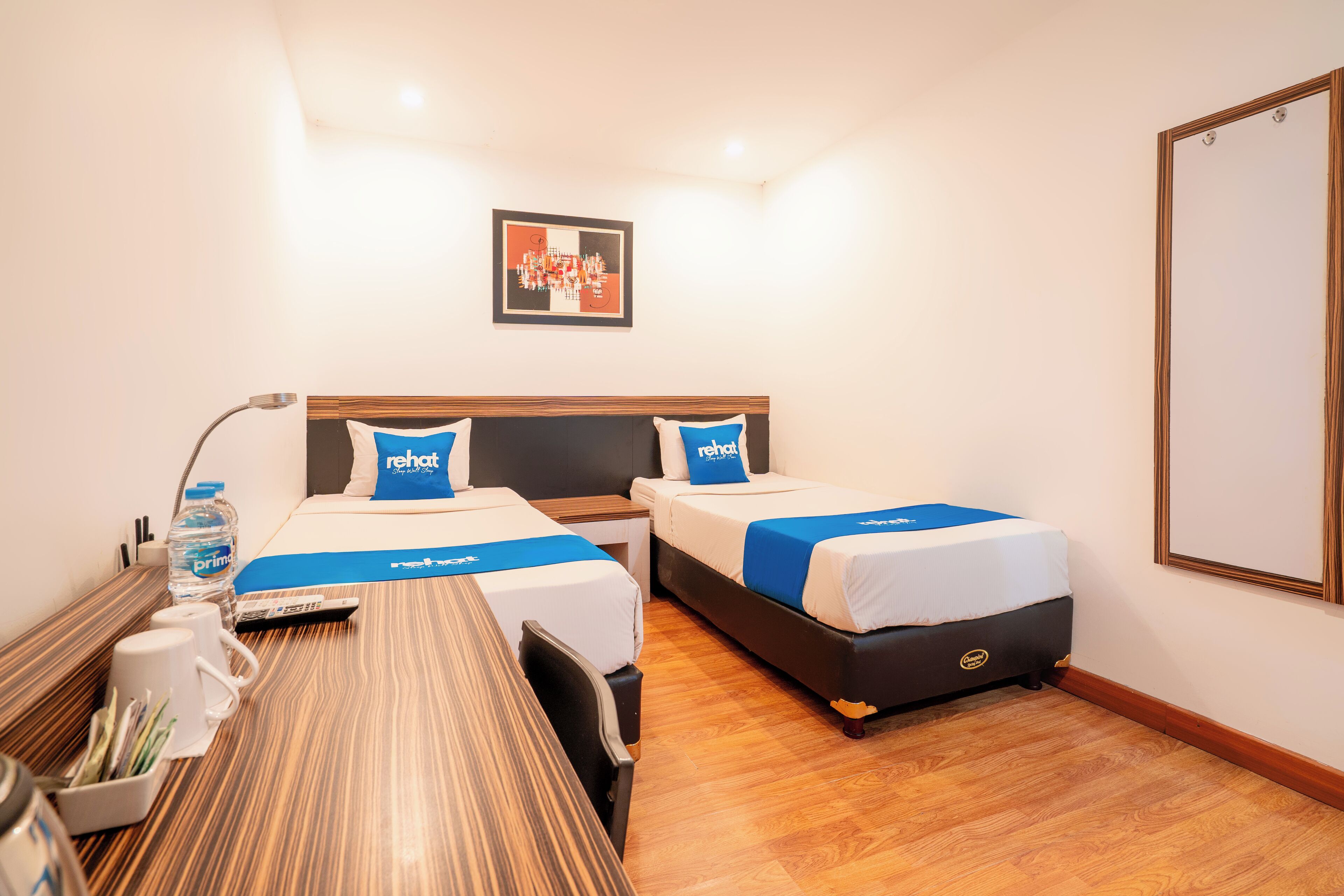A hotel room with two beds, a wooden desk, a chair, a water bottle, a remote control, and a framed picture on the wall.