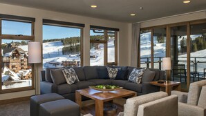 A modern living room with a large sectional sofa, a wooden coffee table, and a view of a snowy mountain landscape through large windows. - ski in/out, amazing 2-bedroom condo in Breckenridge with lots of amenities. (Breckenridge)