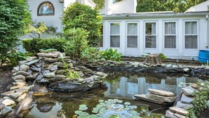 A pond with rocks and plants, surrounded by a white building with large windows. - !Poconos Hidden Villa de’Casablanca Pool+Spa Creek (Stroudsburg)