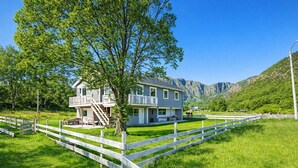A two-story house with a balcony, surrounded by a white fence, set in a green landscape with mountains in the background. - Beach front home in Nordnesøy with house a panoramic view (Nordnesøy)