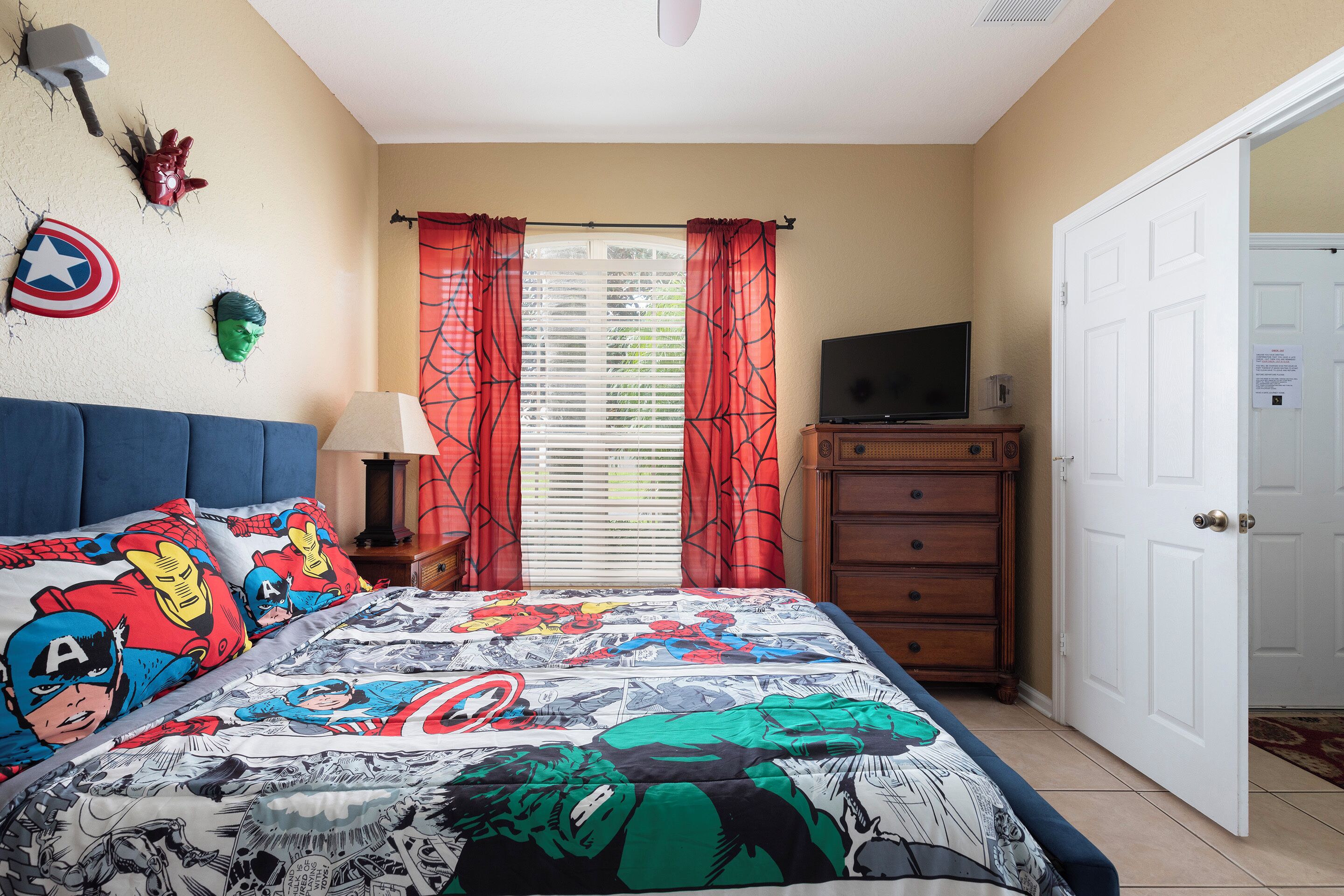 A bedroom with a bed featuring a comic book print, a television on a dresser, and a door to the right.
