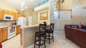 A kitchen with wooden cabinets, a white refrigerator, and a microwave. There are two bar stools with dark seats. A brown sofa is against the wall, and a lamp is on a side table. - Windsor Hills Resort (Kissimmee)