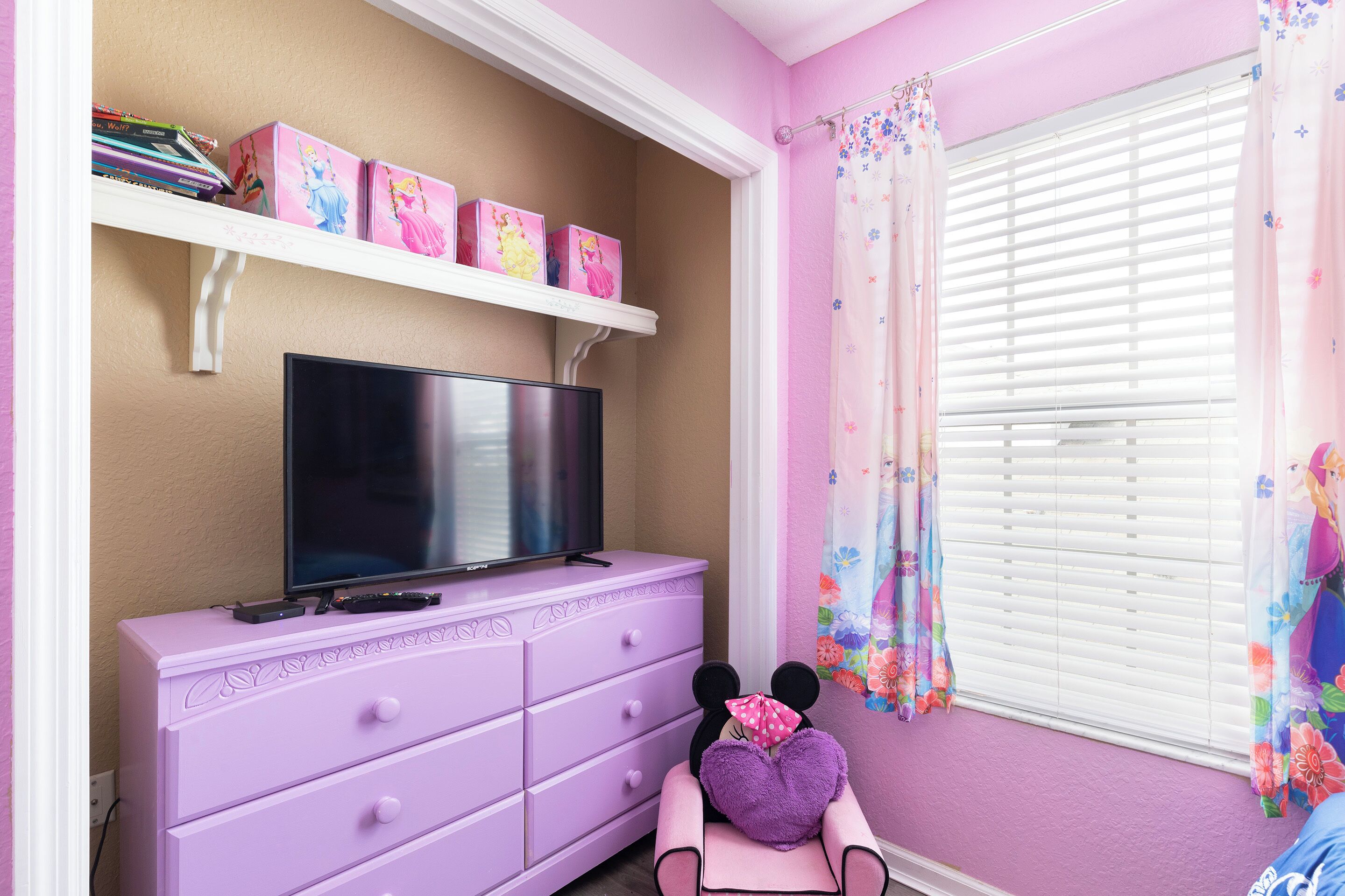 A room with a television on a dresser, a shelf with books and boxes, and a window with floral curtains.