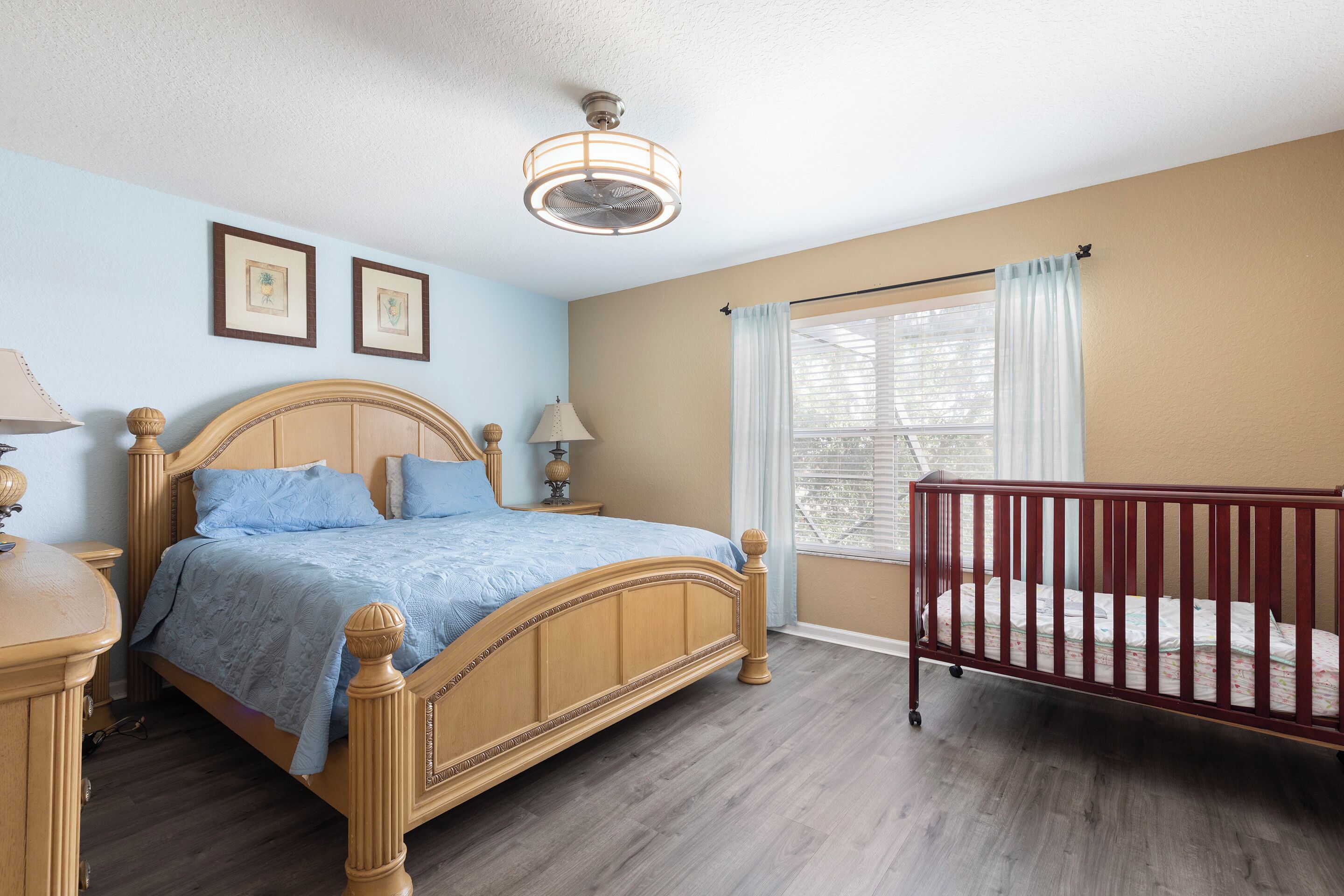 A bedroom with a wooden bed, a crib, a ceiling fan, and a window with curtains.