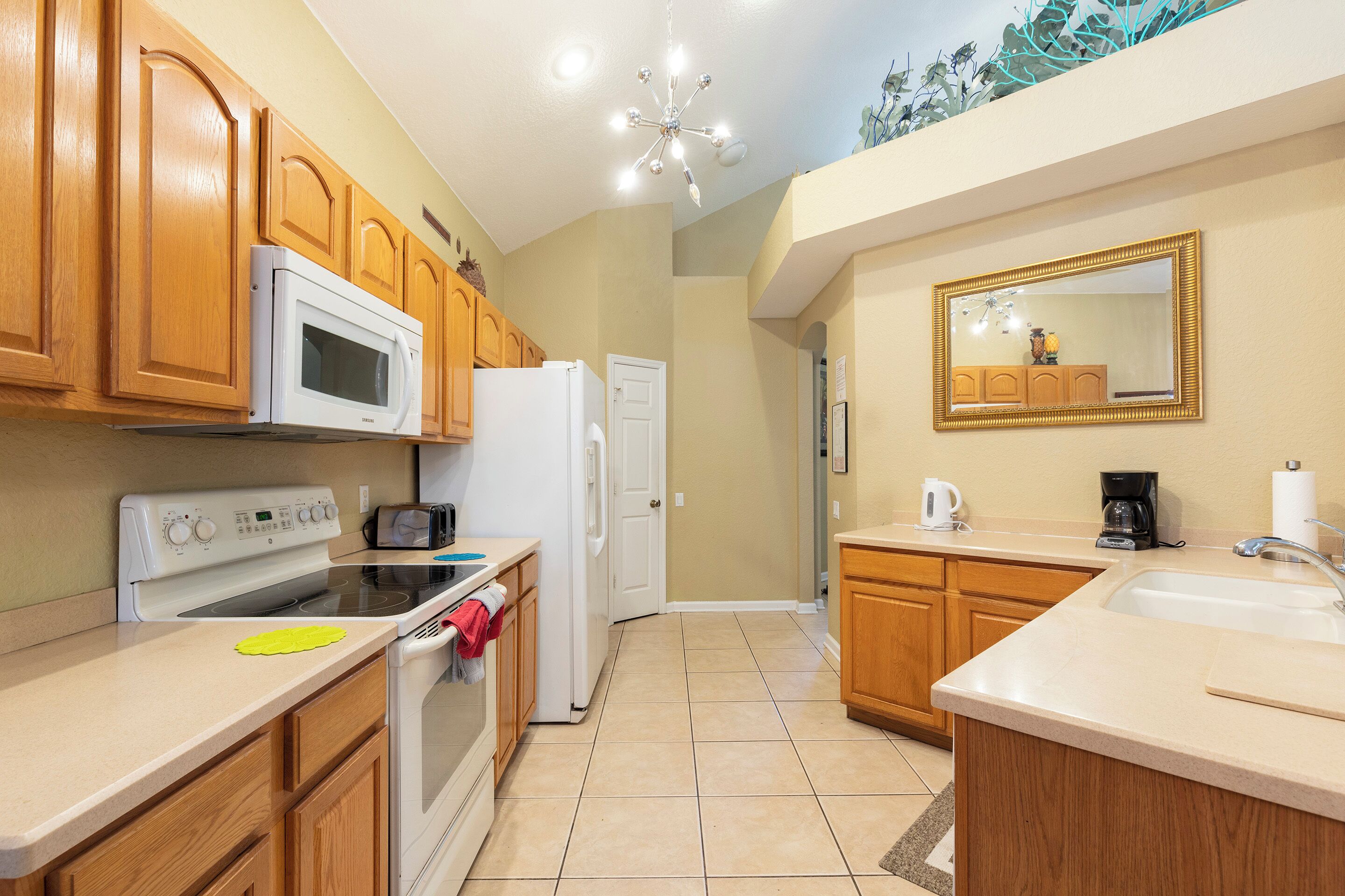 A kitchen with wooden cabinets, a white microwave, stove, and refrigerator. There is a sink and a mirror on the wall.