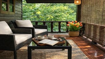 Een rieten bank met kussens, een glazen salontafel met een boek en kopjes, en een potplant met tulpen op een balkon met uitzicht op groen.
