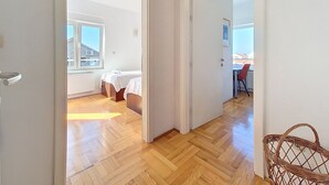 A hallway with wooden flooring, a bed with a white cover, a radiator, and a window with a view of buildings. - Apartment Rilić by Interhome (Zaostrog)