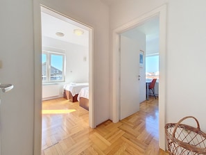 A hallway with wooden flooring, a bed with a white cover, a radiator, and a window with a view of buildings. - Apartment Rilić by Interhome (Zaostrog)